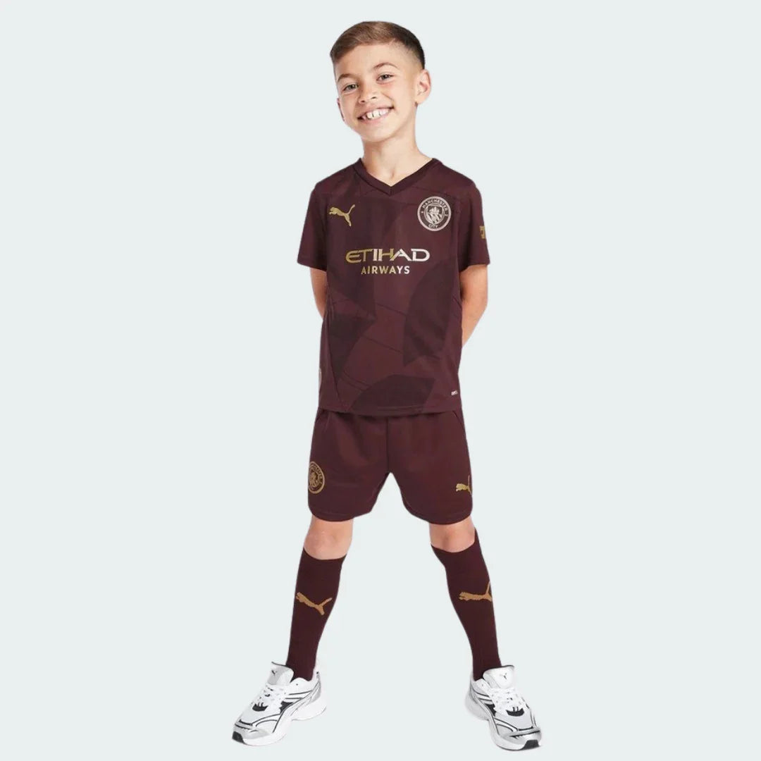 Um jovem garoto sorrindo veste o Kit Infantil Manchester City III 24/25 bordô com detalhes dourados na camisa, shorts e meias.