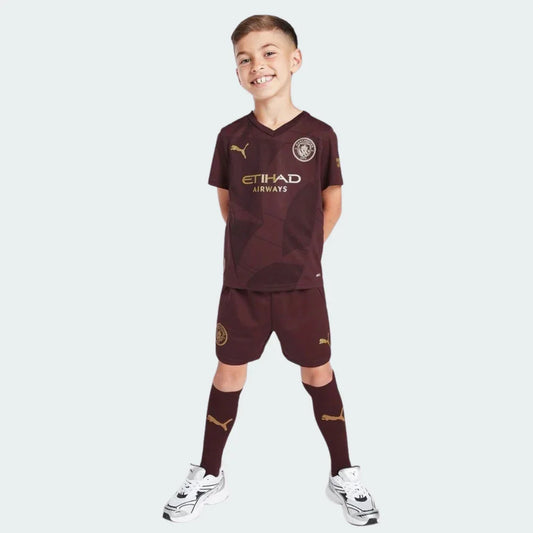 Um jovem garoto sorrindo veste o Kit Infantil Manchester City III 24/25 bordô com detalhes dourados na camisa, shorts e meias.
