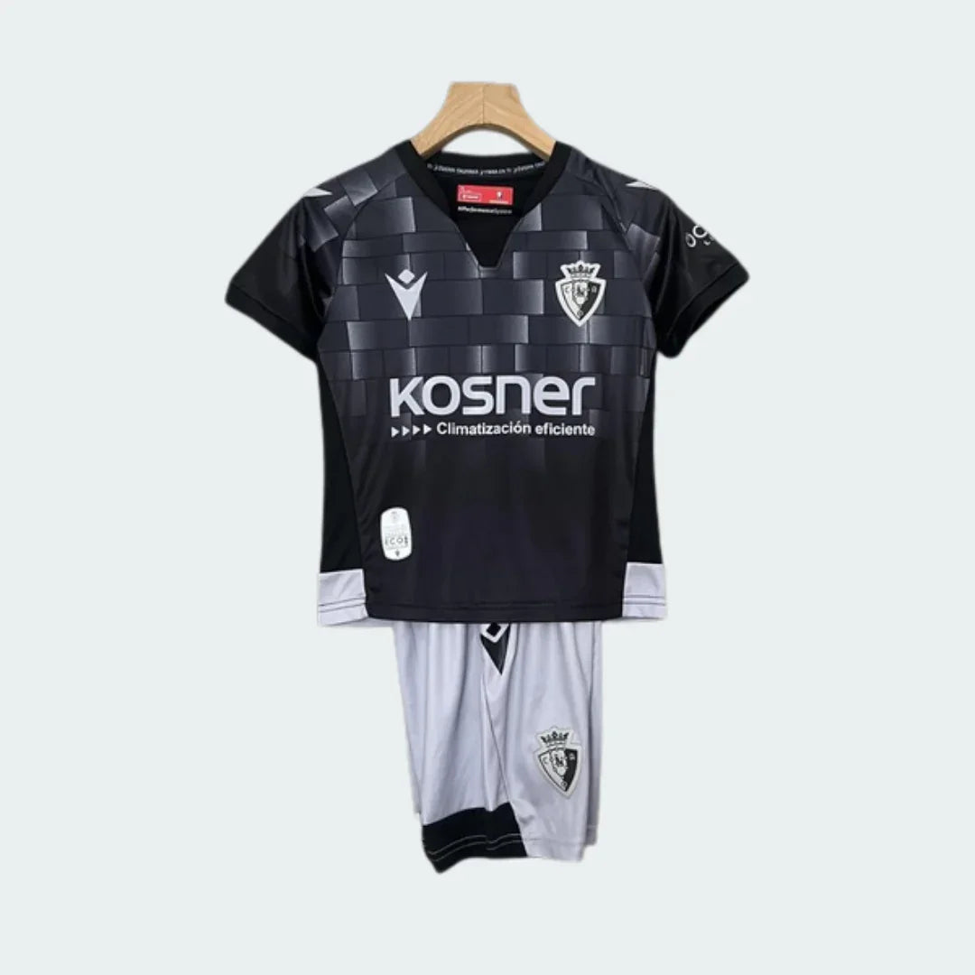 Kit infantil Osasuna III 24/25, camisa preta xadrez com escudo, logo Macron e Kosner. Shorts brancos com escudo do clube, pendurado.
