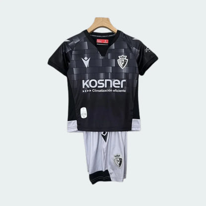 Kit infantil Osasuna III 24/25, camisa preta xadrez com escudo, logo Macron e Kosner. Shorts brancos com escudo do clube, pendurado.