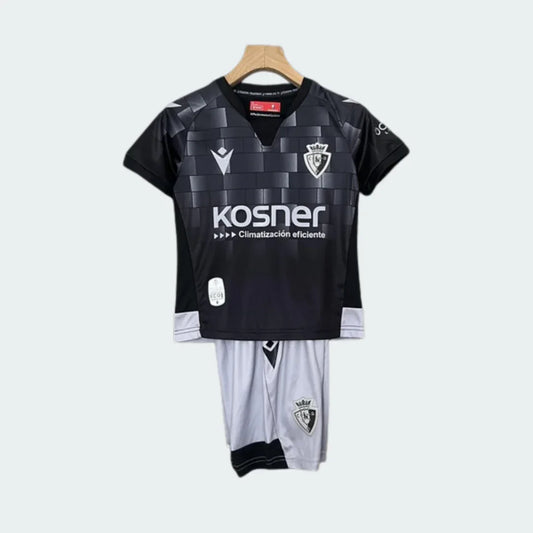 Kit infantil Osasuna III 24/25, camisa preta xadrez com escudo, logo Macron e Kosner. Shorts brancos com escudo do clube, pendurado.