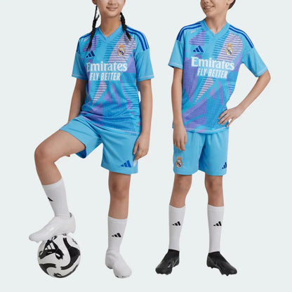 Duas crianças, menino e menina, vestem o kit infantil Real Madrid Portero 24/25 azul claro com estampas roxas e azuis. Um pé na bola.
