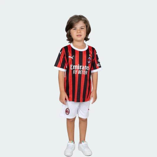 Criança em pé, vestindo kit infantil Milan com camisa listrada preta e vermelha, shorts brancos e tênis brancos em fundo branco.