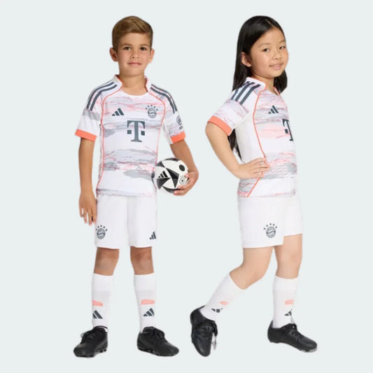 Duas crianças exibem kit Bayern de Munique II 25/26 branco com padrões cinza e laranja claro. Menino com bola, menina sorrindo.
