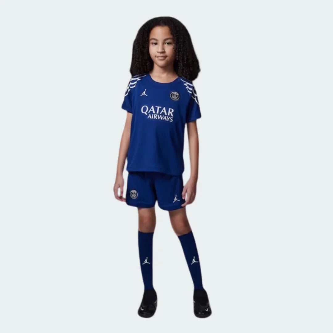 Criança sorrindo vestindo kit infantil PSG azul com detalhes brancos, camisa Qatar Airways, shorts e meias Jordan.