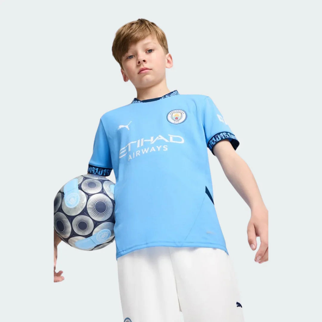 Menino em estúdio vestindo Kit Infantil Manchester City I 24/25 azul celeste, com escudo do time, logo Puma e patrocínio Etihad Airways, segurando bola.
