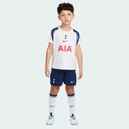Menino em pé, usando kit infantil Tottenham branco e azul marinho. Camisa branca com escudo, logo Nike e AIA vermelho. Shorts azul marinho, meias brancas.