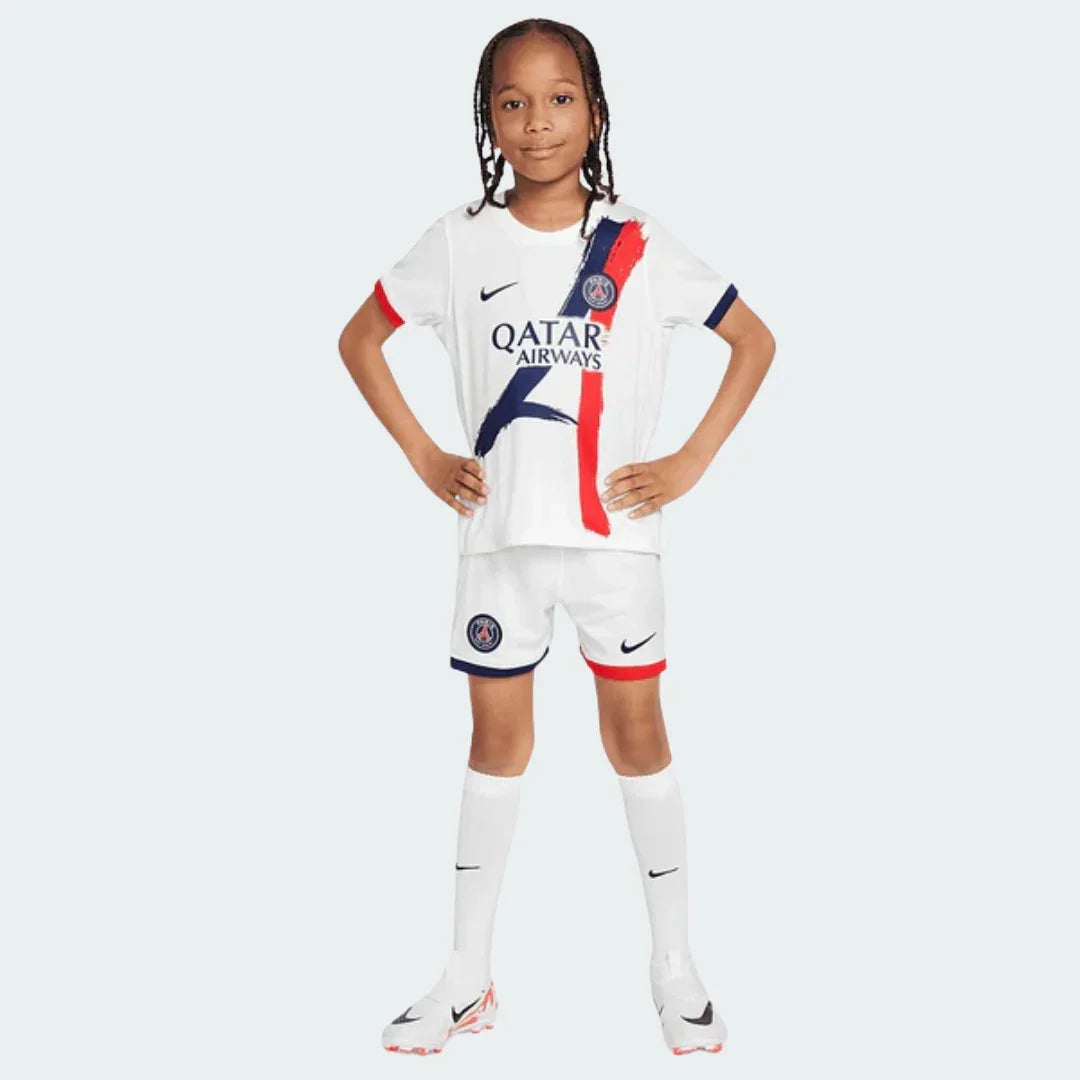 Criança com kit de futebol PSG 24/25 branco, camisa com faixas verticais azuis e vermelhas, shorts e meias. Modelo em pé.