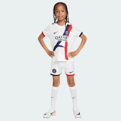 Criança com kit de futebol PSG 24/25 branco, camisa com faixas verticais azuis e vermelhas, shorts e meias. Modelo em pé.