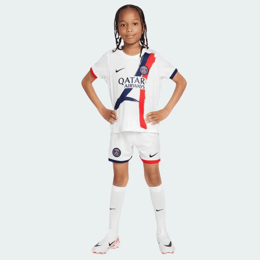 Criança com kit de futebol PSG 24/25 branco, camisa com faixas verticais azuis e vermelhas, shorts e meias. Modelo em pé.