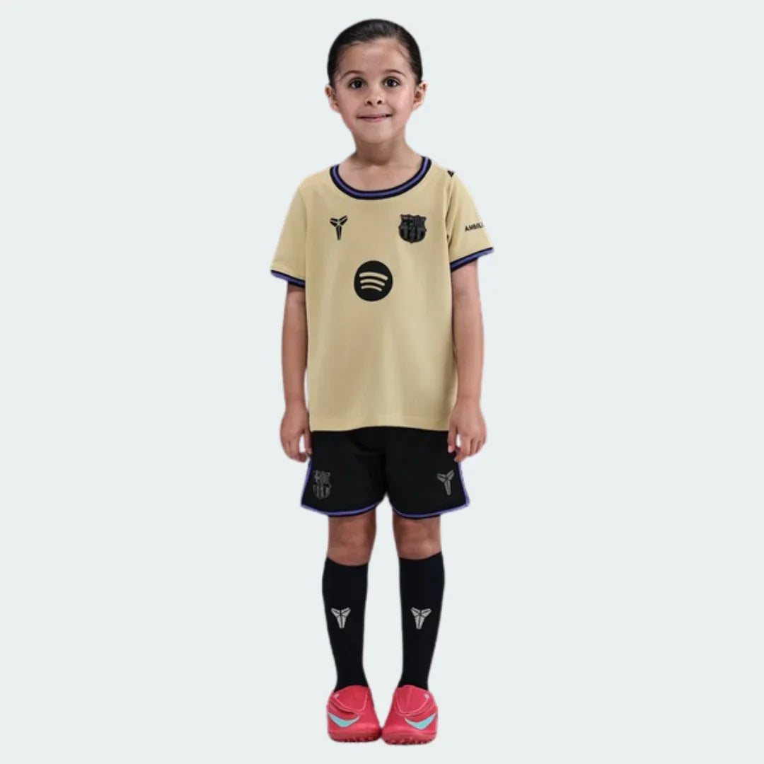 Menina em kit de futebol infantil Barcelona II 25/26, camisa bege, shorts pretos, meias pretas, todos com logos do clube, Spotify e Mamba. Sapatos vermelhos.