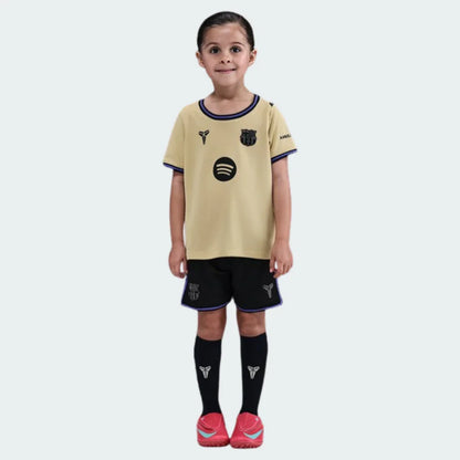 Menina em kit de futebol infantil Barcelona II 25/26, camisa bege, shorts pretos, meias pretas, todos com logos do clube, Spotify e Mamba. Sapatos vermelhos.