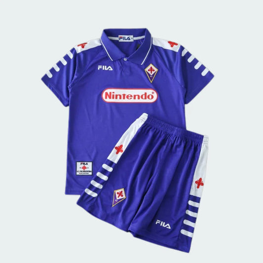 Kit infantil completo da Fiorentina, roxo, com camisa e shorts. A camisa possui gola, logos Fila e Nintendo, além do escudo da Fiorentina. O shorts tem detalhes brancos e vermelhos e o escudo do time.