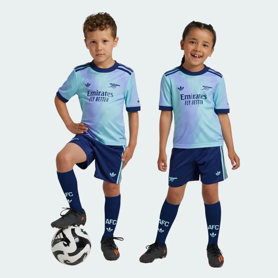Duas crianças sorrindo vestindo o Kit Infantil Arsenal III 24/25, com camisa azul clara e roxa, shorts e meias azul-marinho. Um menino com a bola.