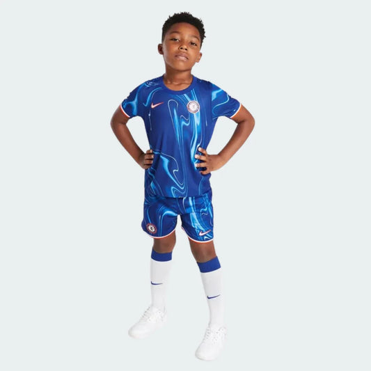 Criança vestindo kit infantil do Chelsea azul com padrão marmorizado, detalhes em vermelho, shorts azuis e meias brancas. Fundo branco.