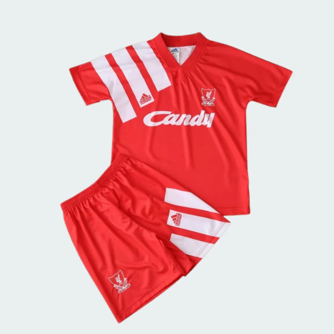 Foto de um conjunto infantil retrô do Liverpool. Camisa vermelha com listras brancas, escudo do clube e patrocínio 'Candy'. Short vermelho com listras brancas e escudo.