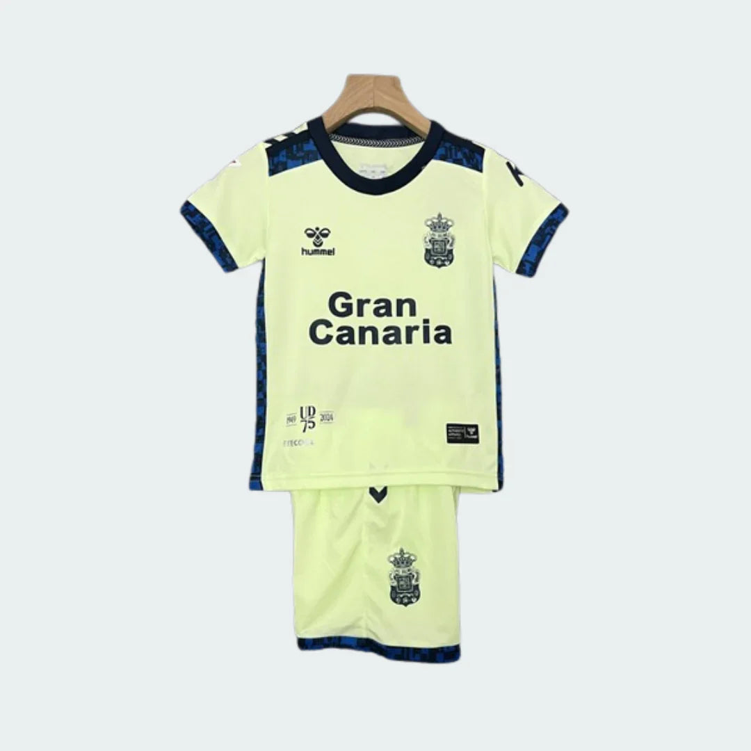 Kit infantil completo do Las Palmas III 24/25, com camisa e shorts amarelos-claros pendurados em um cabide. Detalhes em azul-escuro, escudo, Hummel e 'Gran Canaria' visíveis.