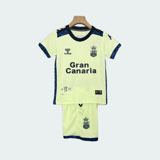 Kit infantil completo do Las Palmas III 24/25, com camisa e shorts amarelos-claros pendurados em um cabide. Detalhes em azul-escuro, escudo, Hummel e 'Gran Canaria' visíveis.
