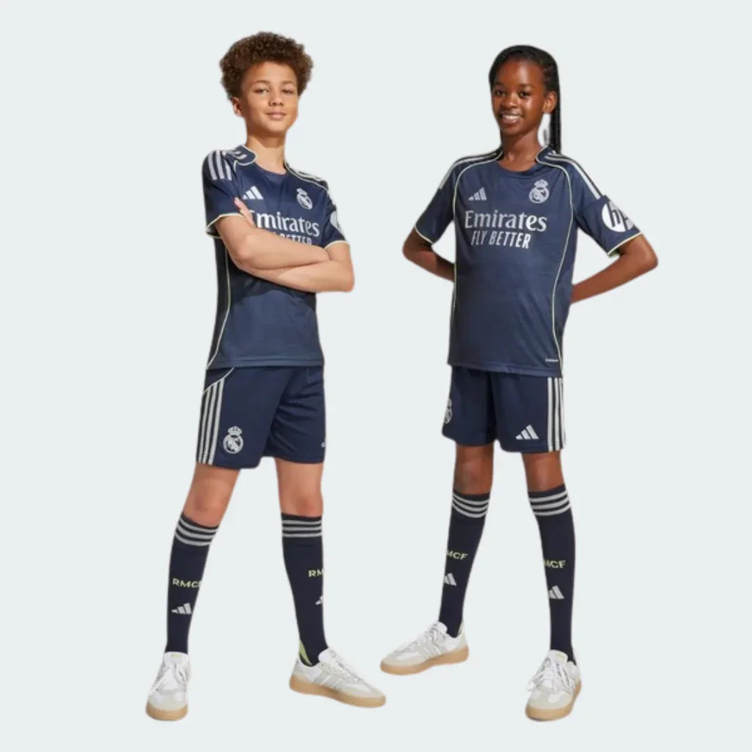 Duas crianças sorrindo vestindo o kit infantil Real Madrid II azul marinho. Camisa e shorts com listras claras, escudo do clube e logo Adidas.