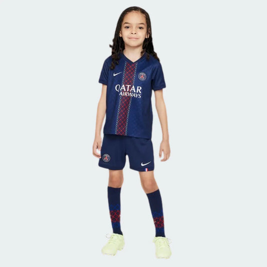 Criança sorridente veste kit infantil PSG I 25/26 azul escuro, com camisa, shorts e meias. A camisa tem faixas verticais vermelhas e brancas.