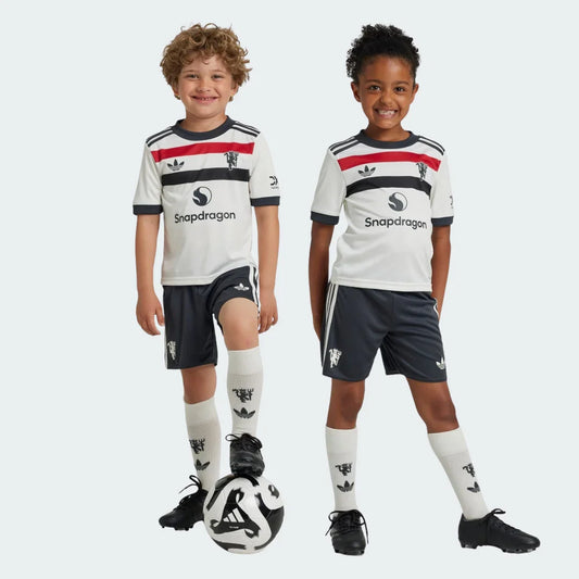 Dois jovens, um menino e uma menina, vestem o kit infantil do Manchester United. Camisas brancas com listras vermelhas e pretas, shorts cinza e meias brancas. Bola de futebol no chão.