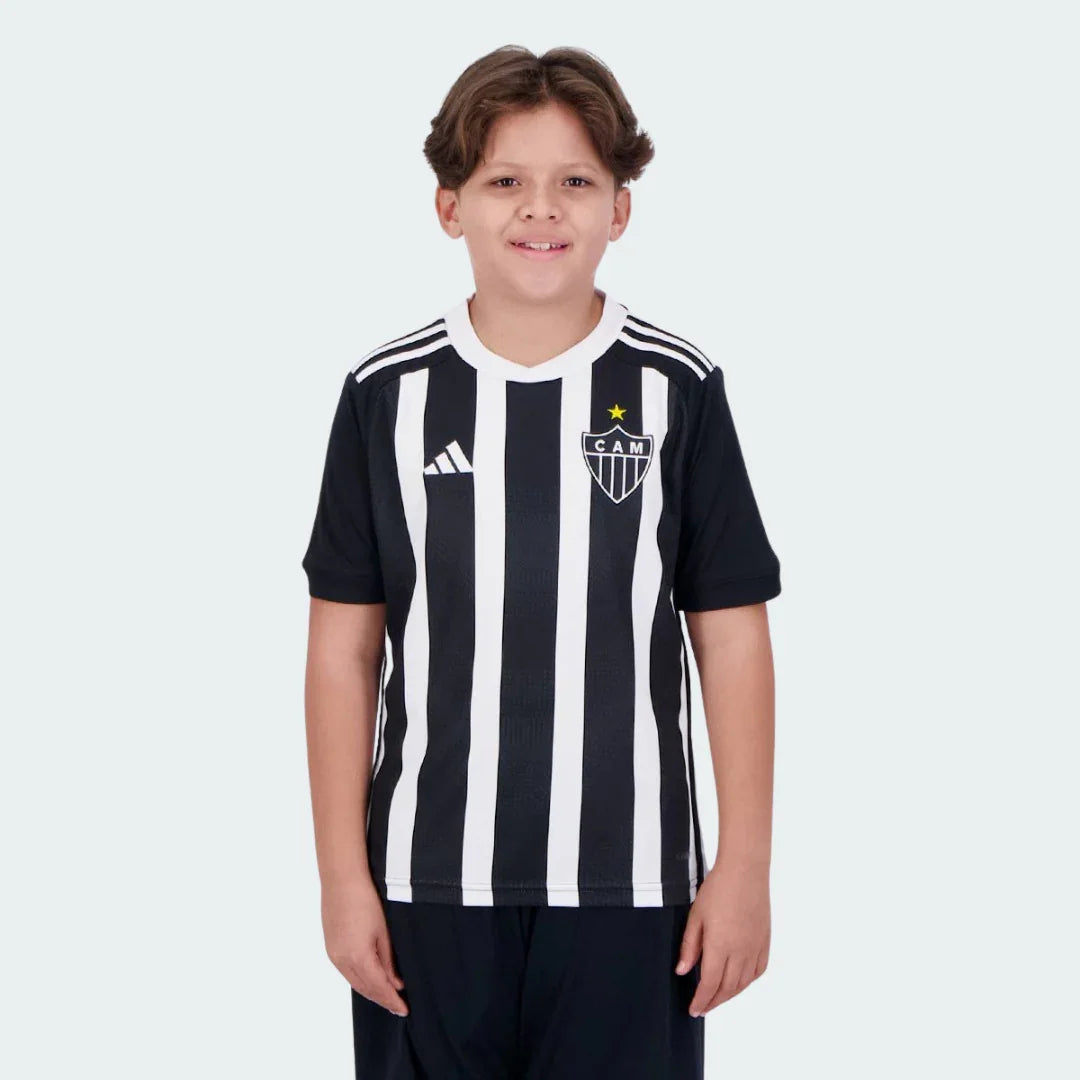 Menino sorrindo veste camisa oficial do Galo I 24/25 preta e branca listrada com escudo CAM, estrela amarela e shorts escuros.