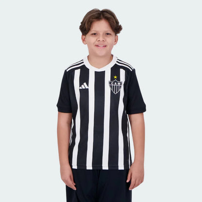Menino sorrindo veste camisa oficial do Galo I 24/25 preta e branca listrada com escudo CAM, estrela amarela e shorts escuros.