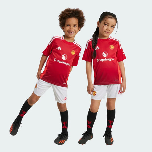 Duas crianças sorrindo em kits infantis do Manchester United: camisa vermelha, shorts brancos e meias pretas com detalhes vermelhos.