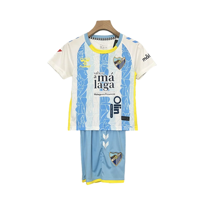 Kit infantil do Málaga 24/25 pendurado, camisa branca e azul com listras abstratas, shorts azuis, detalhes amarelos, escudos do clube.