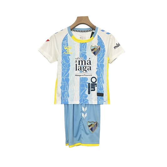 Kit infantil do Málaga 24/25 pendurado, camisa branca e azul com listras abstratas, shorts azuis, detalhes amarelos, escudos do clube.