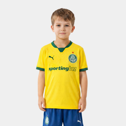 um Menino loiro vestindo o kit infantil do Palmeiras, com camisa amarela, detalhes em verde e calção azul.