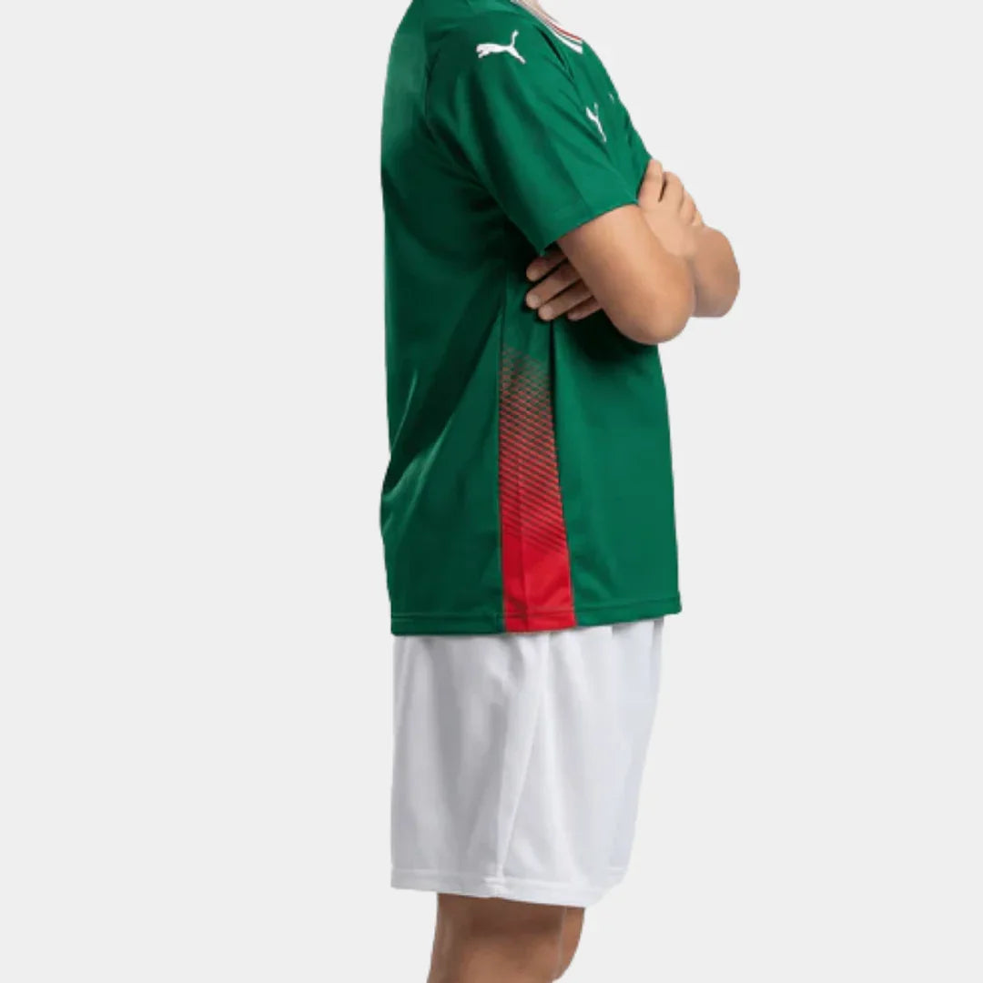 Visão de perfil de um menino com os braços cruzados, destacando o detalhe em mesh vermelho na lateral da camisa verde e o calção branco.