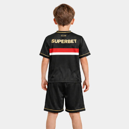 Visão traseira de um menino vestindo o kit completo, destacando a continuidade da faixa tricolor nas costas da camisa preta e o patrocínio "SUPERBET" em letras douradas na parte superior.