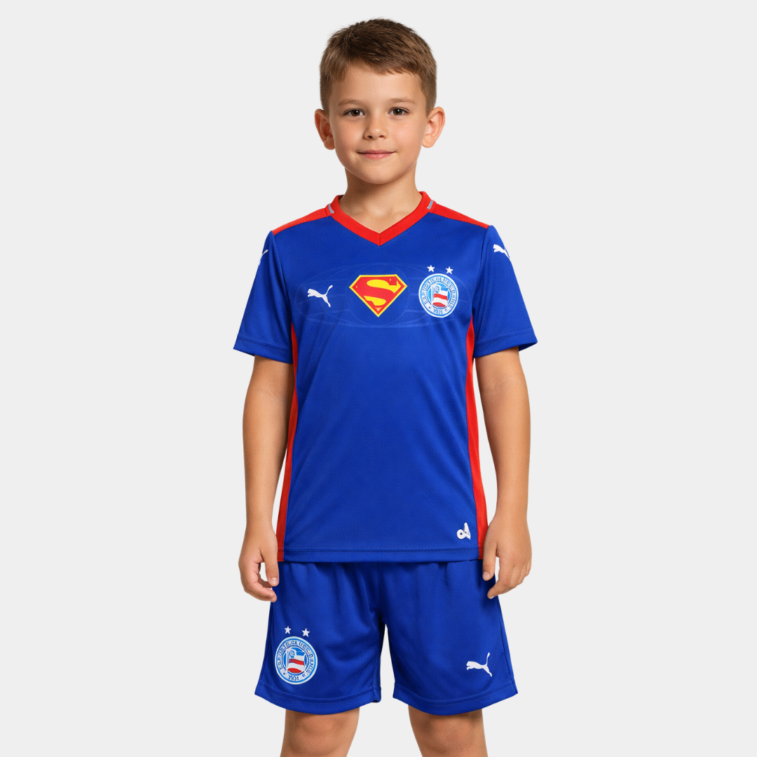 Vista frontal de um menino vestindo o kit infantil do Esporte Clube Bahia na cor azul real, destacando o escudo do Super-Homem centralizado no peito entre o logo da Puma e o escudo do clube, acompanhado de calção azul com os mesmos emblemas.