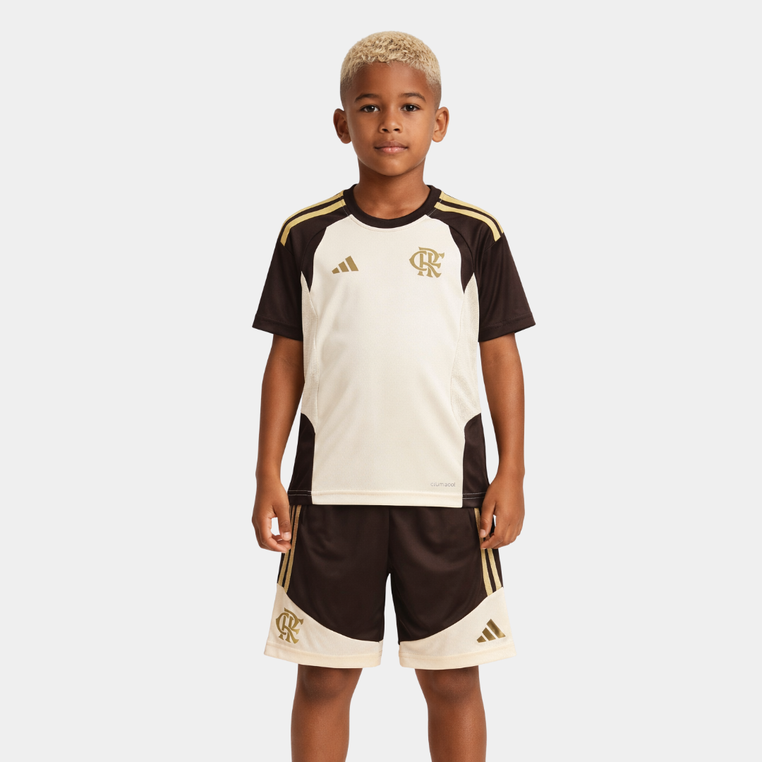 Vista frontal de um menino com cabelo descolorido vestindo o kit infantil reserva do Flamengo, composto por uma camisa off-white com ombros e mangas pretas, detalhes em dourado, acompanhado de calção preto com recortes off-white.