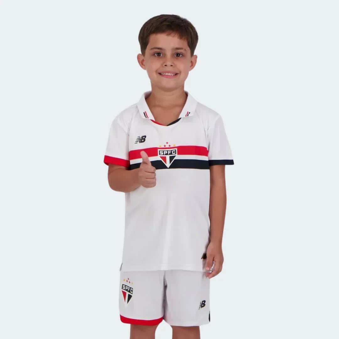 Criança sorridente com kit de futebol branco, com listras horizontais vermelhas e pretas, escudo SPFC e logo NB. Criança faz sinal de positivo.