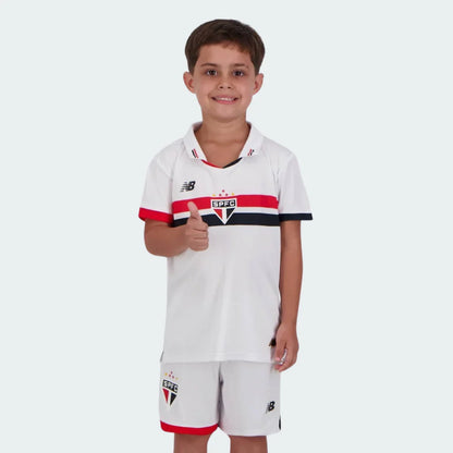 Criança sorridente com kit de futebol branco, com listras horizontais vermelhas e pretas, escudo SPFC e logo NB. Criança faz sinal de positivo.