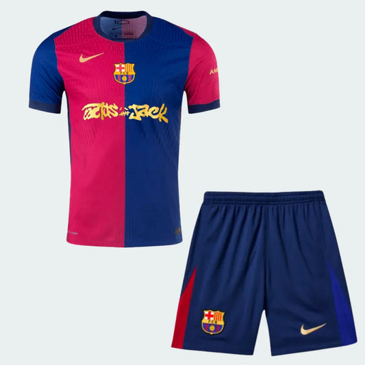 Kit infantil do Barcelona com camisa metade vermelha, metade azul, e shorts azul. Escudo do Barcelona e logos Nike e Cactus Jack.