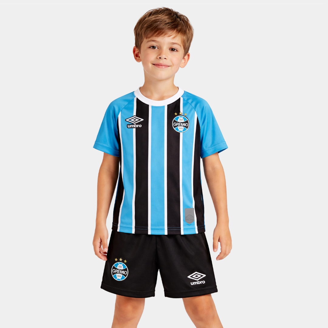 Menino sorridente vestindo o kit infantil titular do Grêmio, apresentando as tradicionais listras verticais em azul celeste, preto e branco, gola careca branca, acompanhado de calção preto com o escudo do clube e o logo da Umbro.