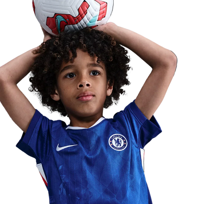 Criança de cabelo cacheado usando camisa azul do Chelsea FC, segurando bola de futebol branca e vermelha acima da cabeça. Fundo preto.