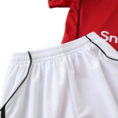 Kit de futebol infantil com camisa vermelha e detalhes brancos, e shorts brancos com detalhes pretos na lateral. Cós elástico.