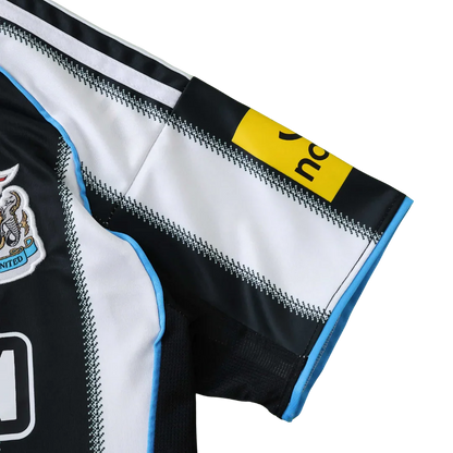 Detalhe da camisa infantil do Newcastle United, listrada em preto e branco, com escudo, detalhes azuis e logo na manga.