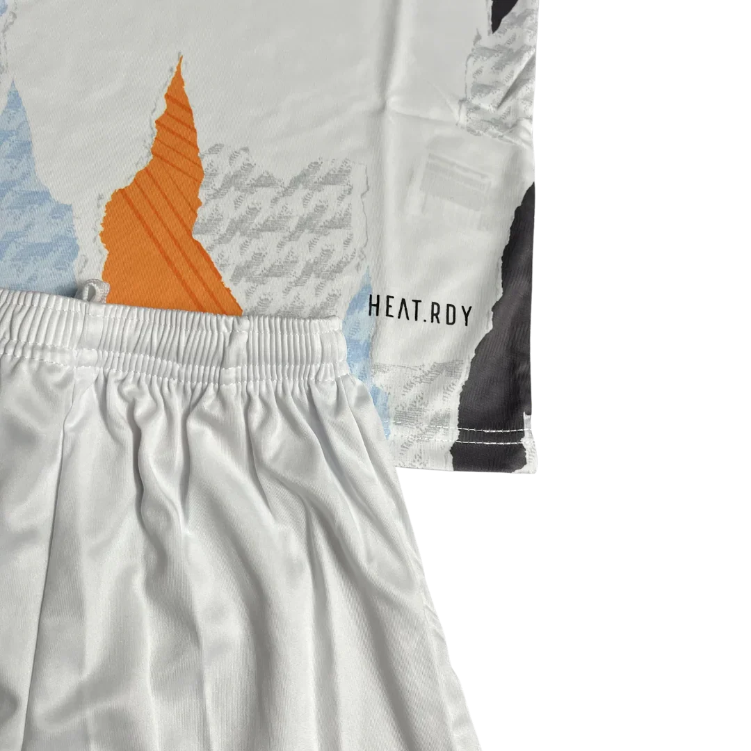 Kit de futebol infantil branco com camisa de estampa abstrata laranja, azul e cinza, e shorts brancos. Detalhe HEAT.RDY visível.