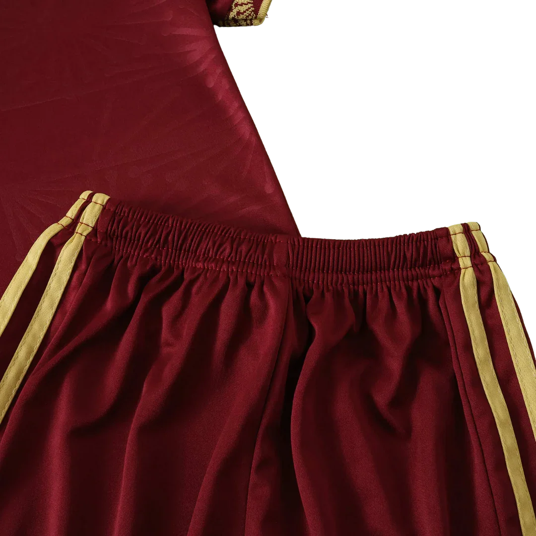 Close-up de shorts de futebol infantil vermelho bordô com cós elástico e listras amarelas. Parte da camisa vermelha ao fundo.