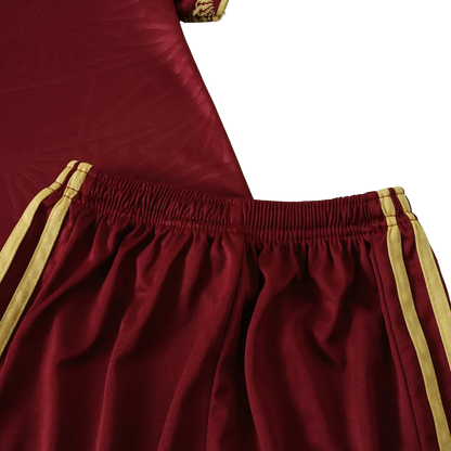 Close-up de shorts de futebol infantil vermelho bordô com cós elástico e listras amarelas. Parte da camisa vermelha ao fundo.