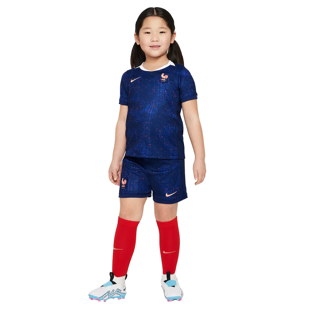 Menina sorridente veste kit infantil da França azul escuro com textura, shorts azuis, meias vermelhas e chuteiras. Posição frontal.