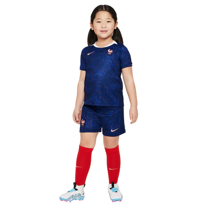 Menina sorridente veste kit infantil da França azul escuro com textura, shorts azuis, meias vermelhas e chuteiras. Posição frontal.