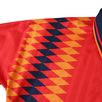Detalhe da camisa de futebol infantil vermelha com padrão diagonal de losangos laranja e azul marinho, e gola azul escura listrada.