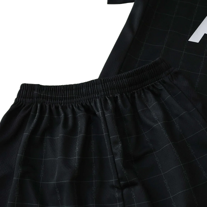 Detalhe de um kit infantil preto, mostrando shorts e parte de uma camisa. O tecido possui padrão quadriculado e texto repetido. Cós elástico e canelado.
