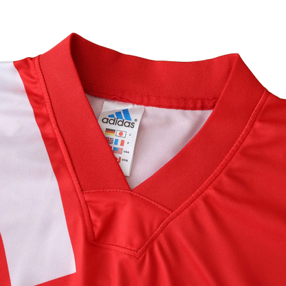 Foto detalhada de uma camisa de futebol infantil vermelha com gola V, listras brancas e etiqueta Adidas visível.